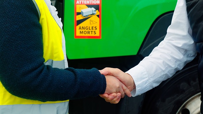 Camion vert en arrière-plan avec deux mains se serrant, symbolisant un partenariat et un accord entre JOST et Renault Lens Motor.