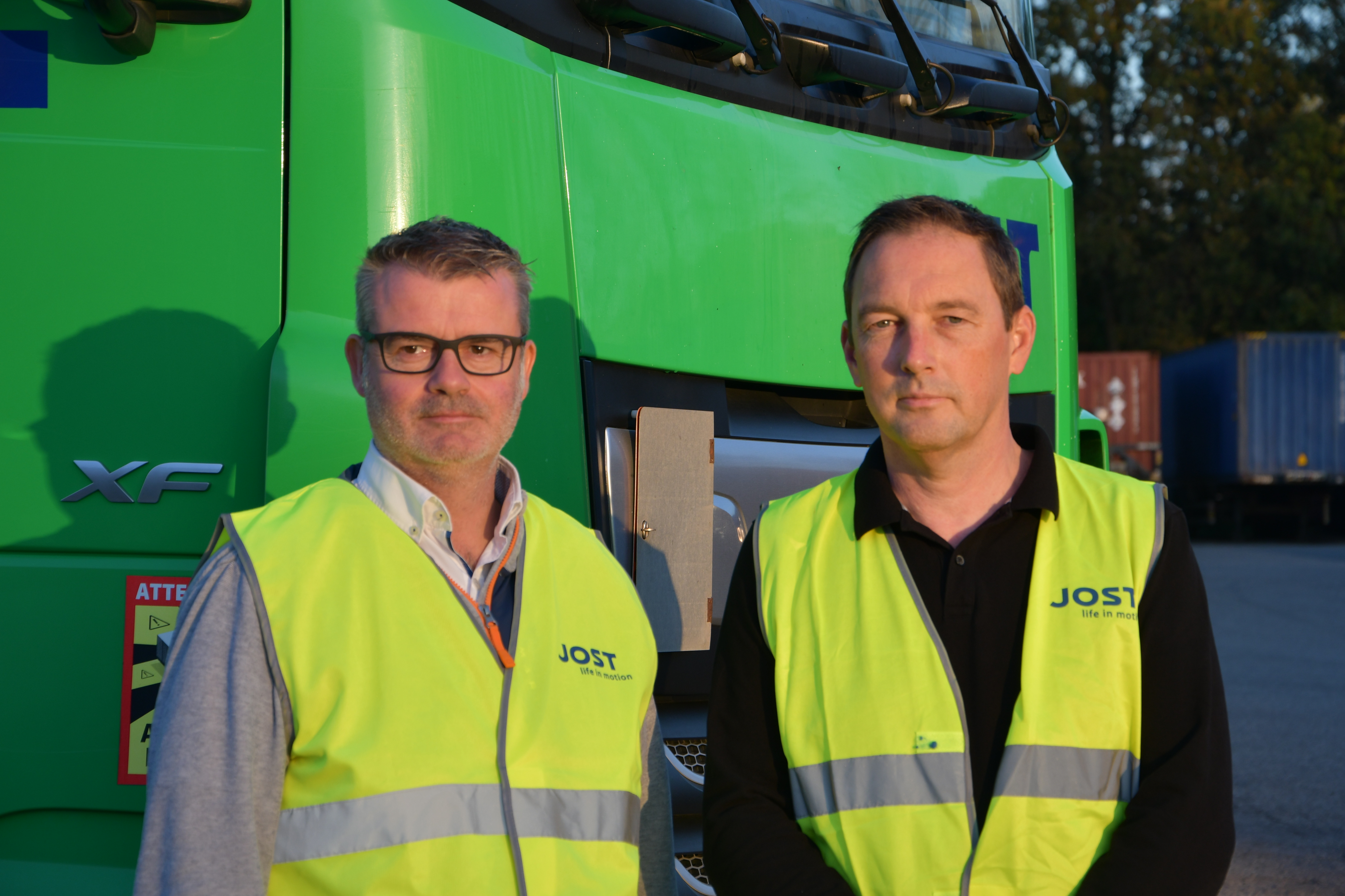 Deux membres de l'entreprise JOST qui posent devant un camion vert JOST avec leur chasuble jaune.