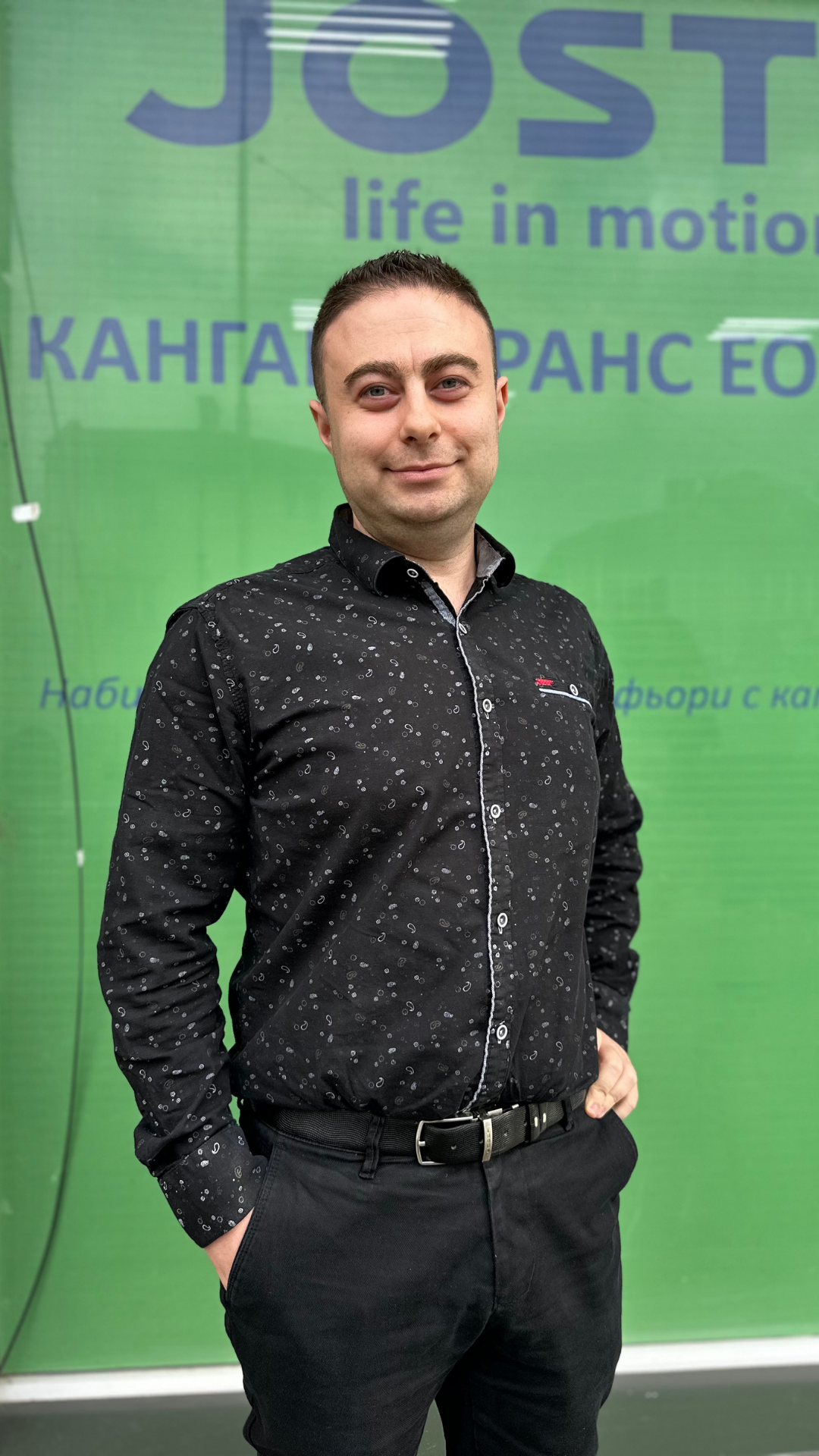 Personne portant une chemise noire à motifs et un pantalon noir, debout devant une vitre verte affichant le logo « JOST life in motion ». La personne est photographiée en pied, avec une main dans la poche, dans un environnement professionnel.