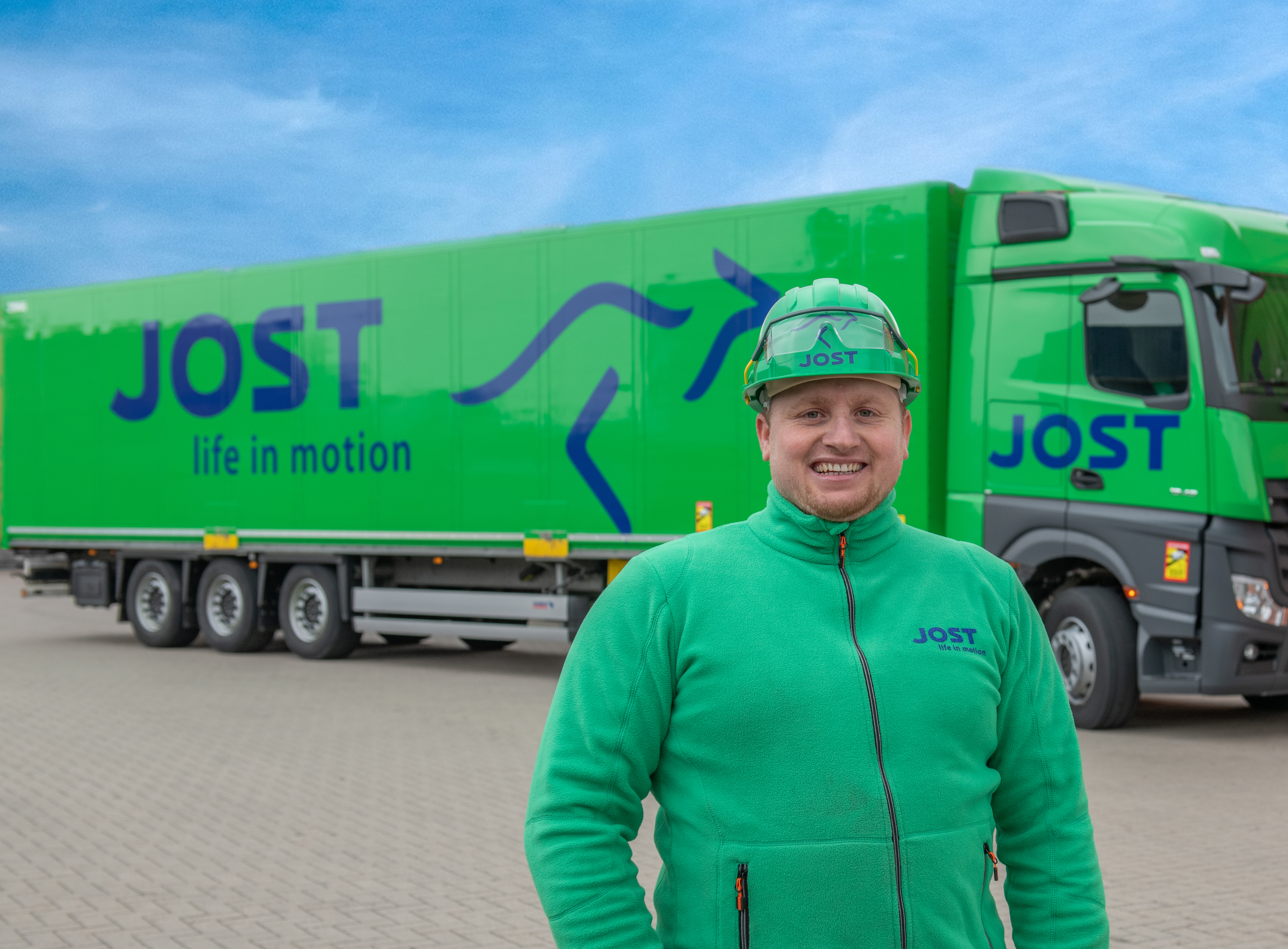 A truck driver wearing a safety helmet stands smiling in front of a Jost transport truck.