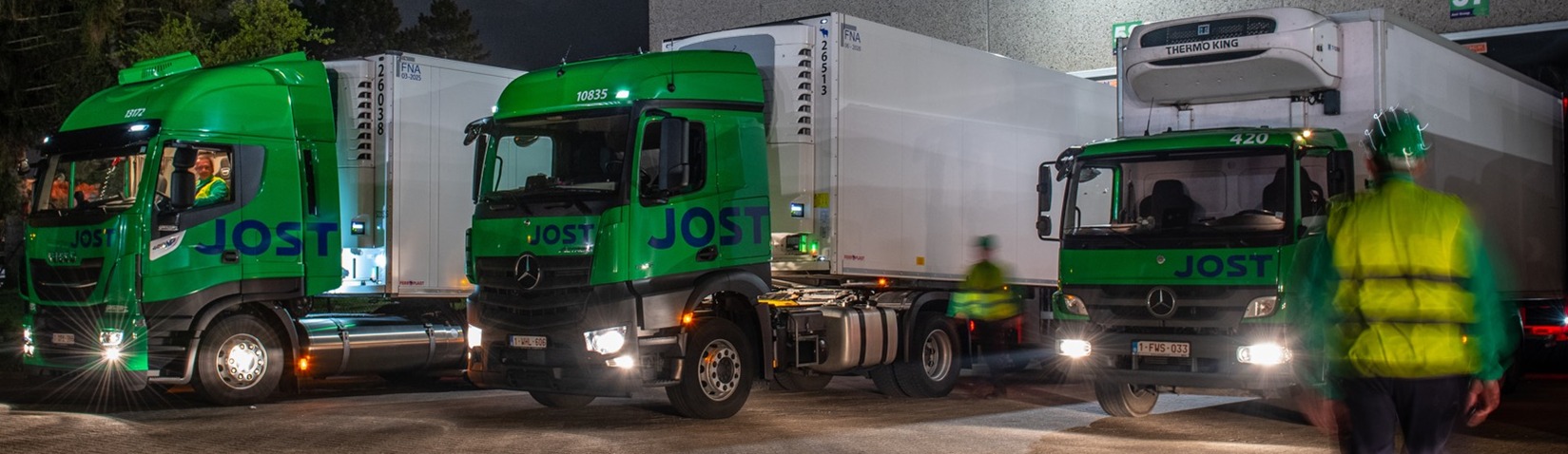 Trois camions de transport JOST devant un hub logistique de nuit avec leurs phares allumés.