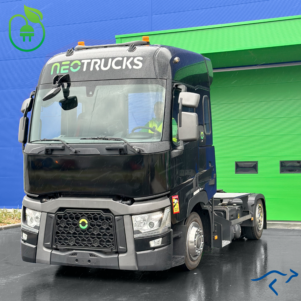 Camion électrique noir devant un bâtiment bleu avec une porte de garage verte sur un tarmac mouillé.