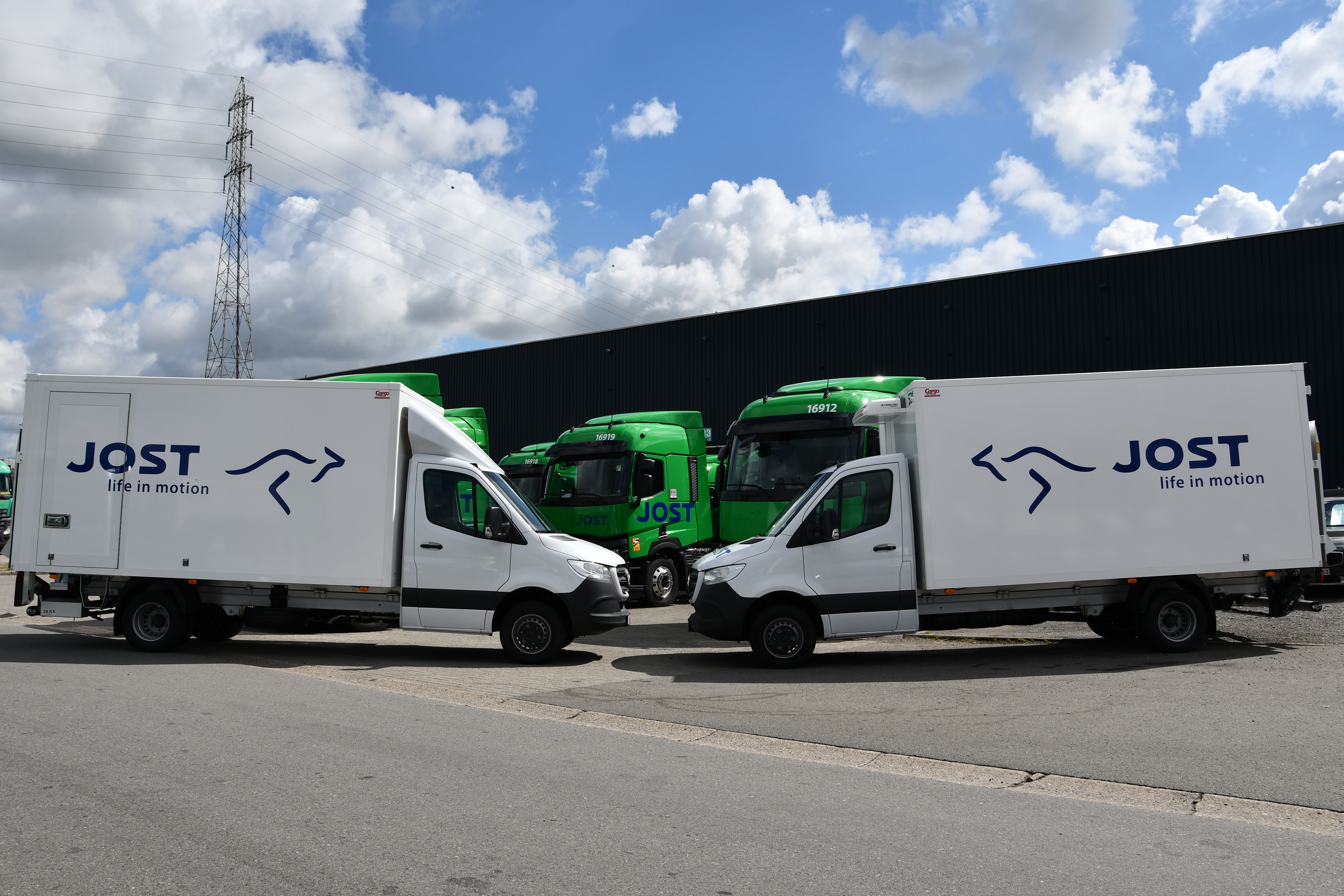 Deux camionnettes blanches de la société de transport Jost garées face à face devant des camions verts.