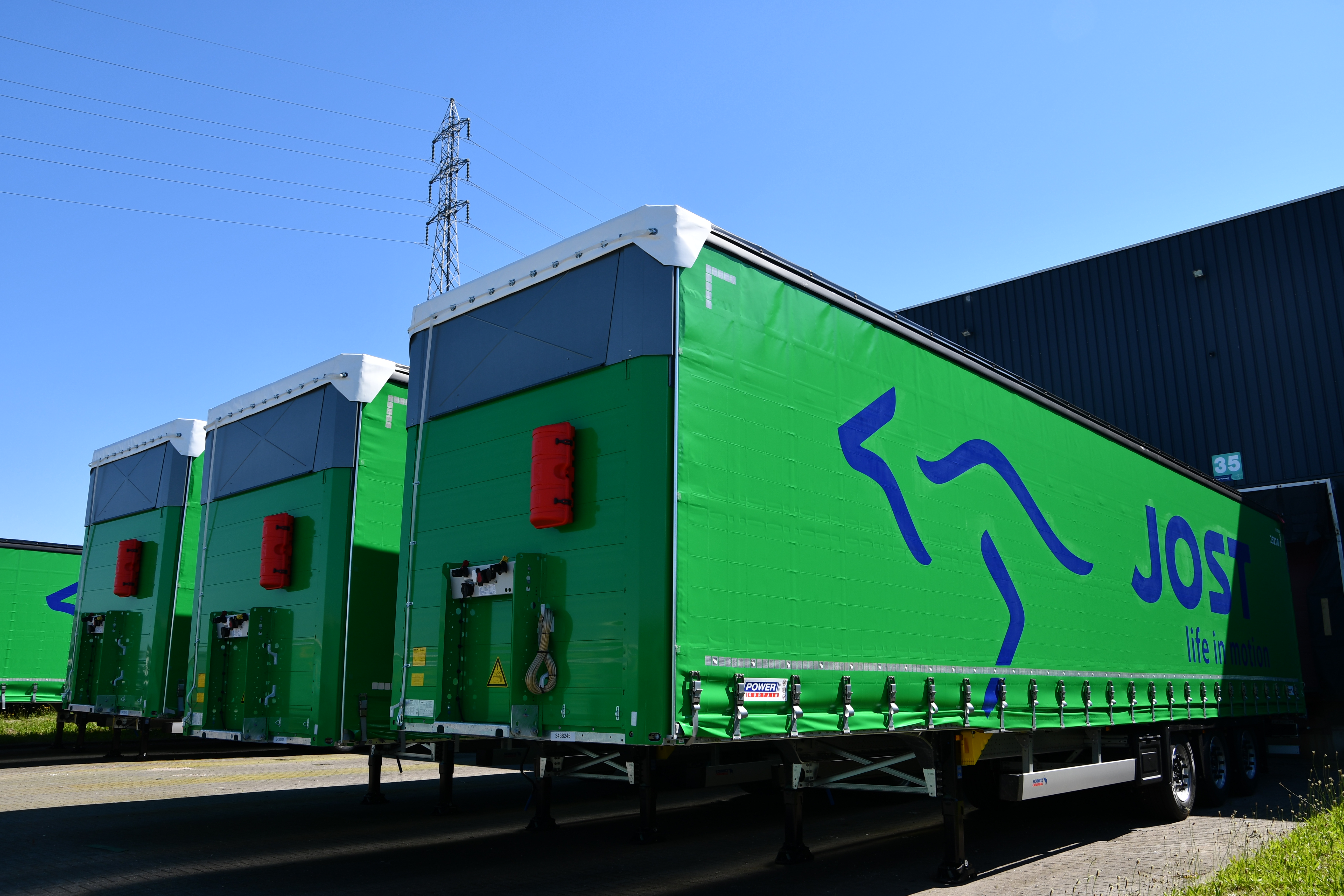 Trois remorques bâchées de la société de transport Jost garées une à côté des autres sans camion avec un ciel bleu.