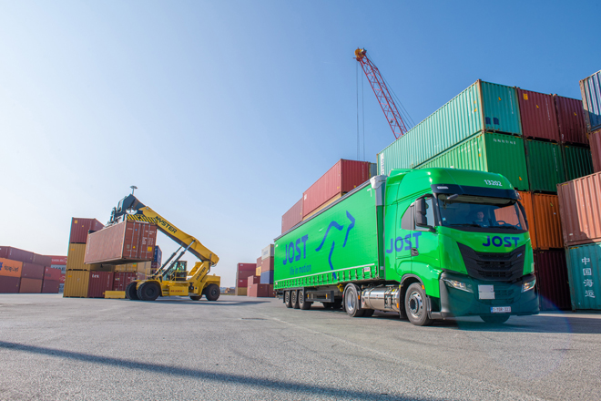 Camion de transport Jost dans un port entourée de conteneurs avec un entrain d’être soulevé par une grue.