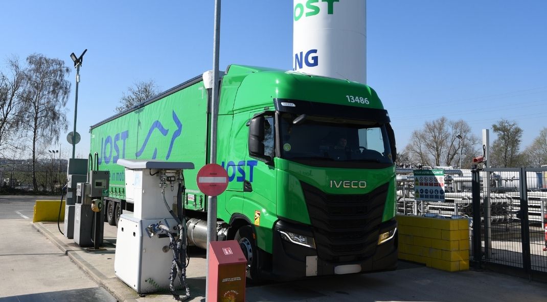 Een groene IVECO-vrachtwagen tankt bij een tankstation, geparkeerd naast een hoge LNG-tank. De omgeving is industrieel, met een heldere lucht en bomen op de achtergrond.