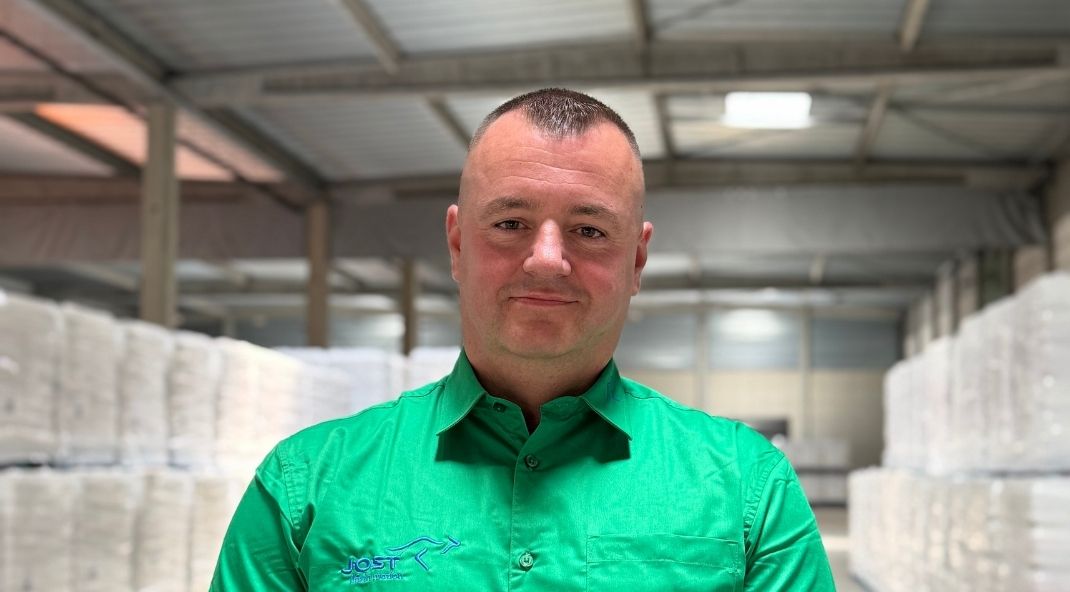 Een man in een felgroen T‑shirt staat zelfverzekerd in een magazijn, omringd door stapels wit verpakte goederen, onder een plafond van metalen balken.