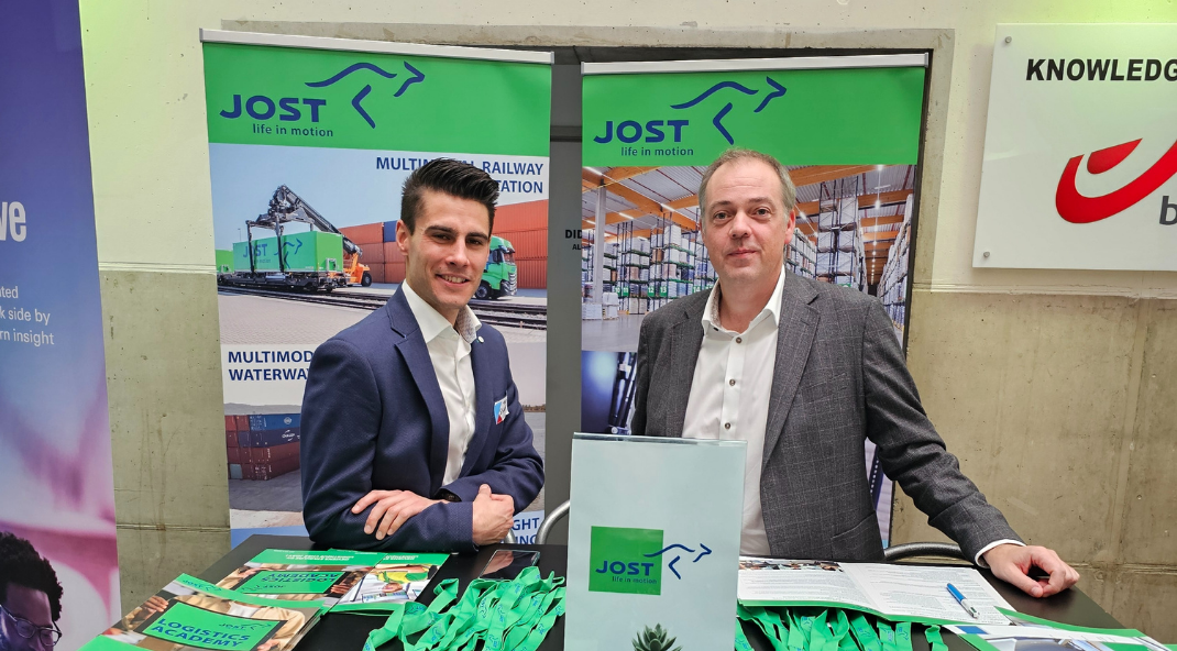 Foto van twee JOST-medewerkers in pak en overhemd, staand voor tafels met goodies en brochures erop.