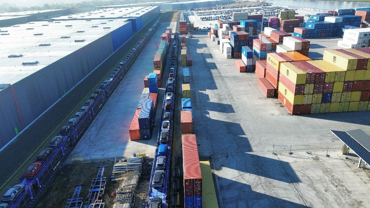 Luchtfoto van een druk industrieterrein met gestapelde zeecontainers aan de rechterkant en beladen treinen aan de linkerkant. De scène straalt efficiëntie en organisatie uit.